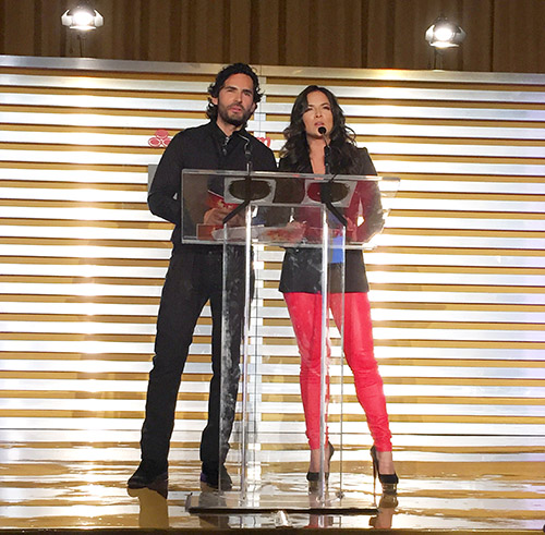 Alt Text: Two presenters speaking at a clear lectern during the 2015 Latin Billboard Awards finalists announcement.