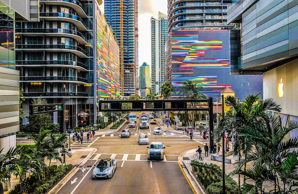 Miami street lined with high-rise buildings, colorful murals, and palm trees, with cars and pedestrians below.