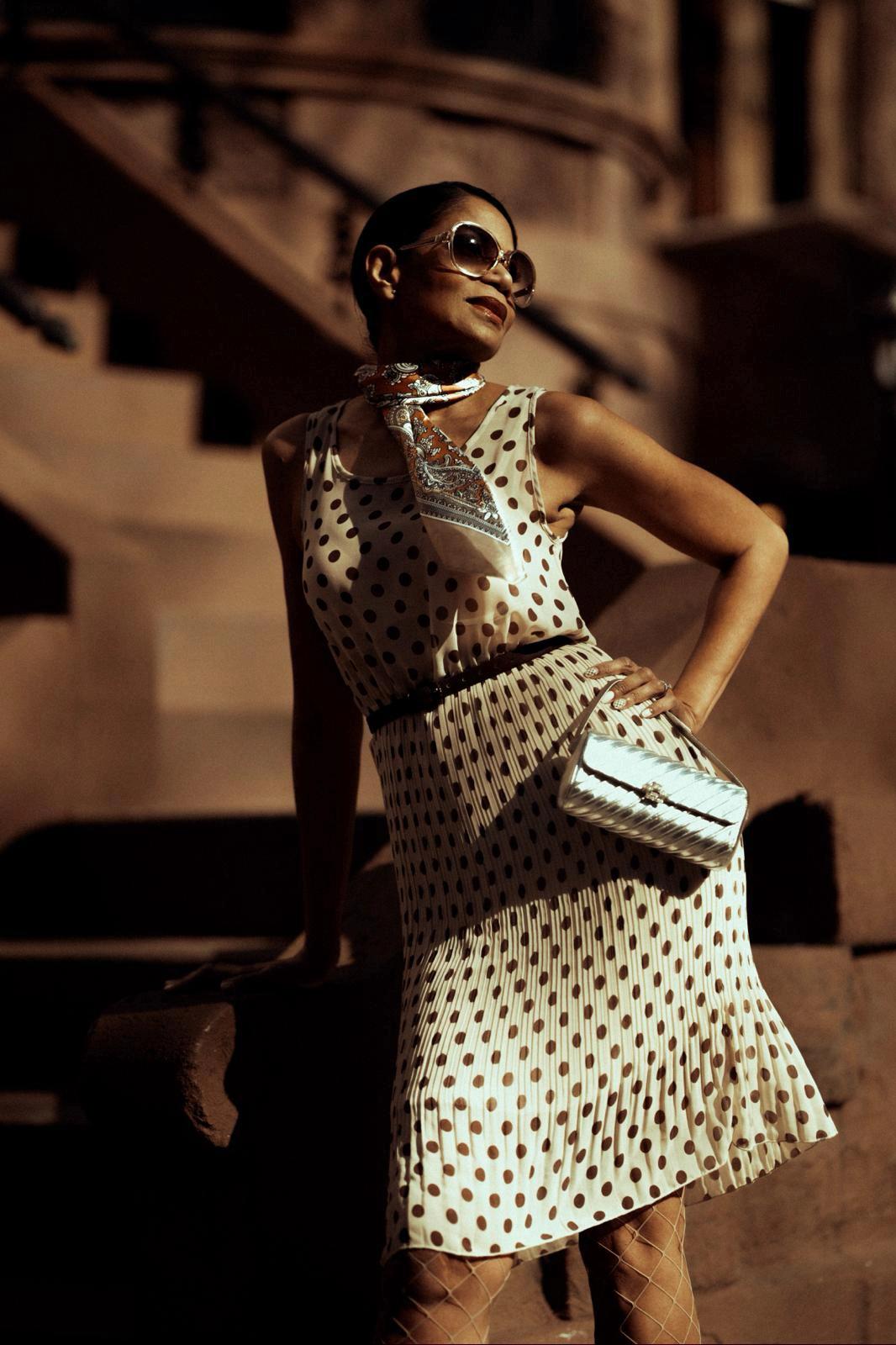 Sandra Ward in a polka-dot dress, sunglasses and scarf, holding a metallic clutch.