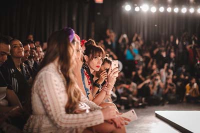 Audience members watching a runway show under stage lights.