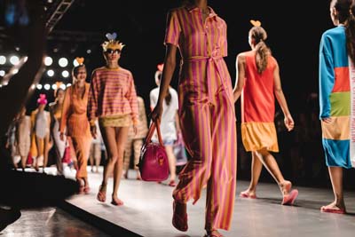 Models walking the runway in colorful dresses and coordinated looks under stage lights.