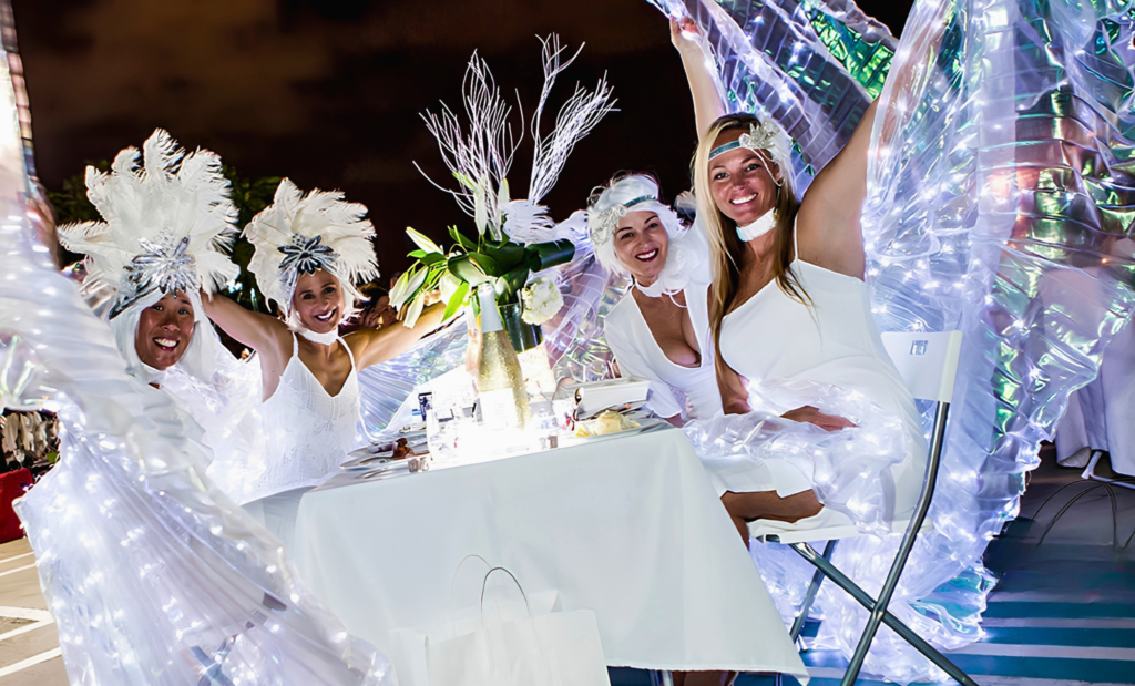 Four guests in white pose beside an outdoor dinner table with feathered headpieces and illuminated wings.
