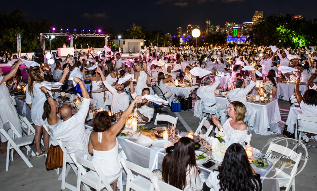 Guests dressed in white wave napkins above outdoor dinner tables at Dîner en Blanc Miami in 2018.
